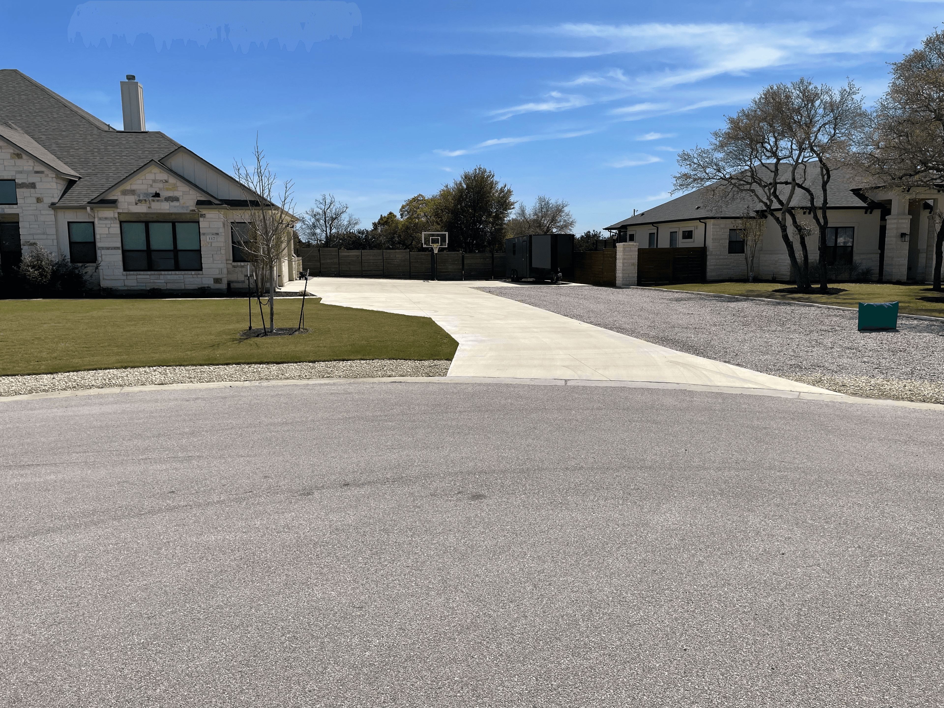 Wide concrete driveway between two stone houses with a basketball hoop in the background.